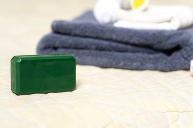 Açık renkli bir yatak örtüsüne yerleştirilmiş yeşil bir sabun. Arka planda düzgünce katlanmış gri havlular ve beyaz tuvalet malzemeleri var..