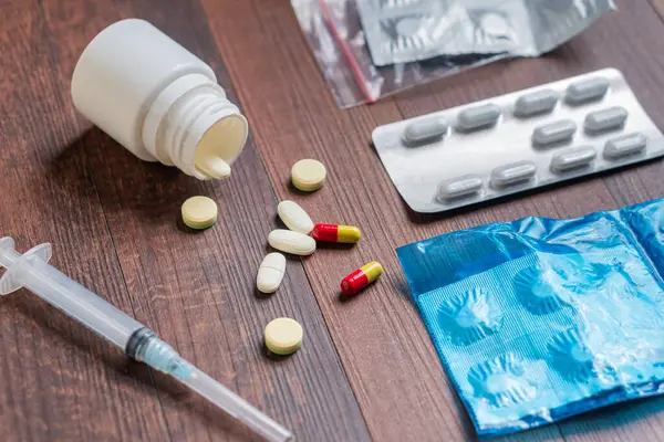 İlaç, şırınga ve tahta bir yüzeyde ilaç şişesi de dahil olmak üzere çeşitli ilaçlar. Sahne, tıbbi bir temayı vurgulayan çeşitli tablet ve kapsülleri tasvir ediyor..