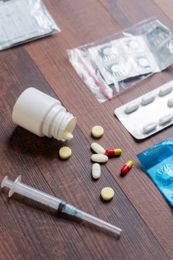 Ahşap bir yüzeyde çeşitli ilaç ve tıbbi malzemelerin toplanması. İçinde dökülmüş bir beyaz hap şişesi, sarı ve kırmızı kapsüller, su toplayan paketler ve bir şırınga var..