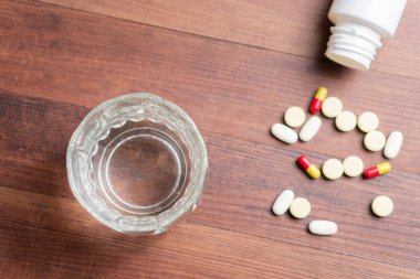 Ahşap bir yüzeyde dağınık hapların yanında temiz bir bardak. Haplar arasında sarı, kırmızı ve beyaz kapsüller var..