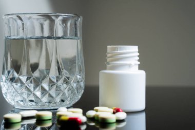 Beyaz ilaç şişesinin yanında bir bardak su ve yansıtıcı bir yüzeye saçılmış haplar..