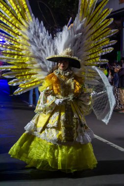 Yogyakarta, Endonezya-6 Ağustos 2025: Tüy kanatlı ve dantel şemsiyeli ayrıntılı sarı bir kostüm giyen zarif bir kadın, canlı bir akşam festivaline katılıyor, yaratıcılık, gelenek ve kutlamalara katılıyor.