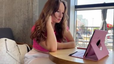 Genç bir kız, genç bir kadın, pembe çantalı bir tabletle bir kafenin penceresinde oturuyor, beyaz eşofman altı, bir çanta ve pembe bir tişört, saçını düzeltiyor, film izliyor, internette vakit geçiriyor, konferans yapıyor.