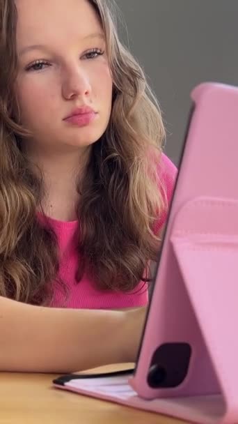 一个年轻的女孩 一个年轻的女人 坐在窗边的咖啡店里 手里拿着粉色的平板电脑 白色的运动裤 一个手提包和一件粉色的T恤衫 直着头发 看电影 在网上消磨时间 开会议 — 图库视频影像