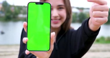Genç, neşeli bir kız gölün arka planında parmaklarıyla gülücükler saçan akıllı bir telefon ekranını işaret ediyor. Yürüyüş eğlencesi için iyi bir reklam. Açık tenli kız.