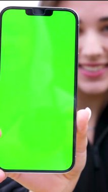 Genç, neşeli bir kız gölün arka planında parmaklarıyla gülücükler saçan akıllı bir telefon ekranını işaret ediyor. Yürüyüş eğlencesi için iyi bir reklam. Açık tenli kız.