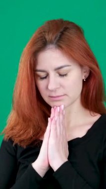 dikey video. Krom anahtarlı bir stüdyoda yeşil arka planda genç, kızıl saçlı bir kadın. Ağız kenarında el ele dua eden beyaz bir kadın, kendinden emin hissediyor. Kanada.