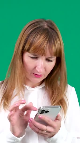 クロマキースタジオで緑の背景に携帯電話の若い女性にメッセージを入力するベージュシャツのコカシアのビジネスマンの傾きのサイドスローモは 光肌のヨーロッパのアメリカ人を閉じる感情を閉じます カナダ — ストック動画