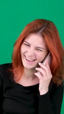 dikey video. Krom anahtar stüdyo duygularında yeşil arka planda olan kızıl saçlı genç bir kadın. Kahverengi gözlü, açık tenli, Avrupalı Amerikalı. Kızıl saçlı kadın telefonda yüksek sesle gülerek konuşuyor. 