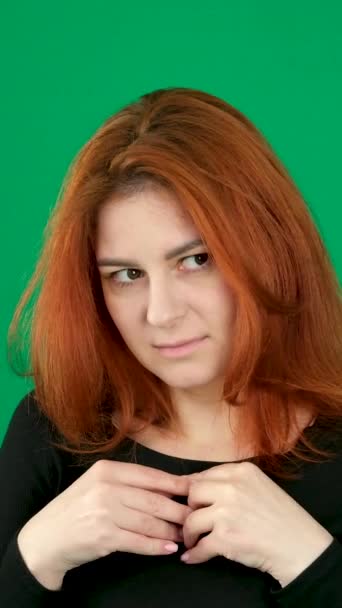 垂直ビデオで クロマキースタジオの緑色の背景にある赤い髪の若い女性は 恥ずかしがり屋の指の下のまつげは 胸の期待に失敗した手を折ります ホープライトスマイル — ストック動画