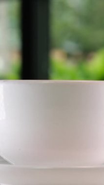 Masanın üzerinde beyaz seramik bardak bulanık arka planda yakın plan, pürüzsüz parallaks. Yaz terasında sabah kahvesi.