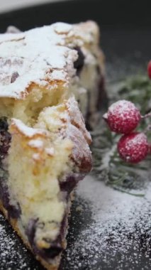 Yılbaşı ağacı süslemeleri ve noel ağacı süslemeleri ile birlikte siyah bir tabak noel yemeği Rowan üzümü üzerinde kameranın önünde dönen lezzetli meyveli turta. Üzerine pudra şekeri serpilmiş.