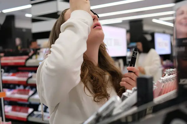 cosmetics shop In store, sample of lipstick is chosen for lip gloss, young girl chooses lip gloss near mirror. buys cosmetics puts on makeup with backlight beauty adolescence teenage girl. woman.