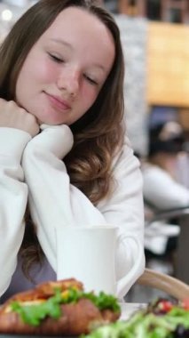 Genç bir kadın bir kafe restoranında kahvaltı yapıyor. Yanında kruvasan ve siyah tabakta salata var. Hoş duygular, gülümseme, seyahat etmenin keyfi, siyah tabakla yalnız kalmak..