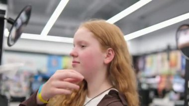 Mağazada dudak parlatıcısı için ruj numunesi seçiliyor, genç kız aynanın yanında dudak parlatıcısı seçiyor. Kozmetik ürünleri alır arka plan ışıklarıyla makyaj yapar ergenlik çağındaki genç bir kızla. Kadın.