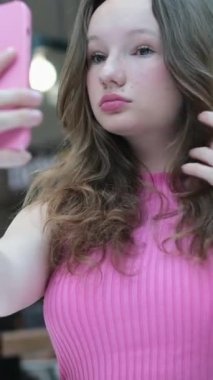 Genç bir kız kafeteryada kendi fotoğrafını çekiyor, saçını düzleştiriyor, telefona bakıyor, sosyal ağlar aracılığıyla arkadaşlarına fotoğraf gönderiyor, onları internete, pembe kapağa yüklüyor. pembe ceket