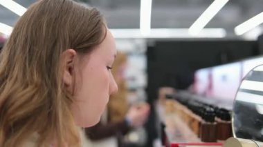 Mağazada dudak parlatıcısı için ruj numunesi seçiliyor, genç kız aynanın yanında dudak parlatıcısı seçiyor. Kozmetik ürünleri alır arka plan ışıklarıyla makyaj yapar ergenlik çağındaki genç bir kızla. Kadın.