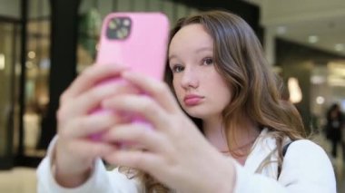 Alışveriş merkezindeki genç bir kız selfie çeker, saçını düzeltir, kendi fotoğraflarını çeker, göz yapar, dudaklarını büzer, gülümser, ergenlik çağında, kamera önünde gösteriş yapar, kendini filme çeker, kısa videolar çeker. Yüksek kalite