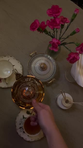 クラフトアンティークテーブルウェアゴールデンガラスティーポットで飲むお茶 女性がピンクのカーネーションの美しい花の背景に対してカップに紅茶を手渡すアジアの香りが近くに立っています — ストック動画