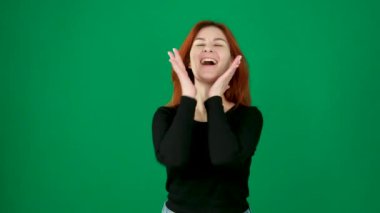 Siyah elbiseli kızıl saçlı kız neşeyle gülümsüyor, mutlu bir şekilde, elleriyle yüzüne sarılıyor, sonra avuçlarını birleştiriyor duygular, kızıl saçlı genç kadın yeşil arka planda, yakın plan krom bir anahtarda. 