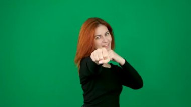 Siyah elbiseli kızıl saçlı bir kız boksör duruşu içinde durur, gülümser ve yumruğunu sallar. Yeşil arka planda kızıl saçlı, yeşil arka planda genç bir kadın. Krom anahtar stüdyo duygularında 4k yakın plan.