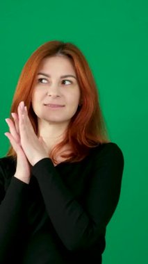 Siyah elbiseli kızıl saçlı bir kız, mutlu ve mutlu, avuçlarıyla ellerini ovuşturuyor, gülümsüyor. dikey video. Krom anahtar stüdyo duygularında yeşil arka planda olan kızıl saçlı genç bir kadın. 