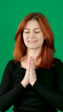 Siyah elbiseli kızıl saçlı kız gülümsüyor ve meditasyon yapıyor, dua ediyor, avuçlarını birleştiriyor, farklı duygular içinde. Krom anahtarlı bir stüdyoda, yeşil arka planda genç bir kadın dikey video çekiyor.