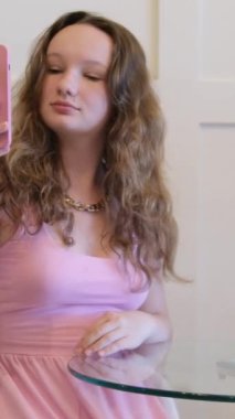 Pembe elbiseli bir kız, cam bir masada, bir kafede, arka planda beyaz bir duvar ve palmiye yapraklarıyla selfie çekiyor. Yüksek kalite 4k görüntü