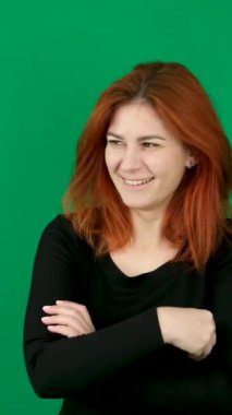 Kızıl saçlı kız gülümsüyor, kollarını göğsünde kavuşturuyor ve başını eğiyor. dikey video. Kızıl saçlı genç bir kadın yeşil arka planda Krom anahtar duygular yakın kahverengi gözler açık ten 