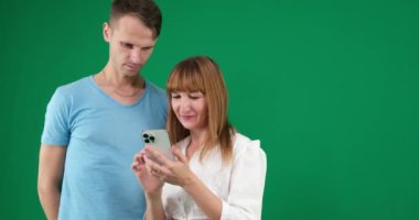 Genç bir çiftin izole edilmiş yeşil arka planda poz vererek cep telefonundan komik videolar izlediği bir video. Yüksek kalite 4k görüntü
