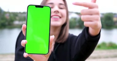 Gülümseyen bir kız işaret parmağını yeşil ekranlı akıllı telefon ekranına doğrultuyor. Reklamcılık. Yüksek kalite 4k görüntü