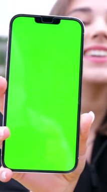 Gülümseyen bir kız işaret parmağını yeşil ekranlı akıllı telefon ekranına doğrultuyor. Reklamcılık. Dikey videolar, yüksek kalite 4k görüntü