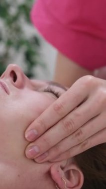 Profesyonel yüz masajı, tapınaklar ve kulaklar, güzellik salonunda yüz masajı yapan kadın. Dikey video