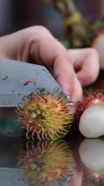 Kadın eli, Rambutan 'ı bıçakla soyar. Cam bir masada egzotik meyveler. Ağır çekim. Dikey video, yüksek kaliteli FullHD görüntüler