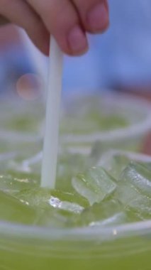 Plastik bardakta buzla pipetle şeker kamışı içen genç bir kız içeceği karıştırıyor. Dikey video, yüksek kaliteli FullHD görüntüler