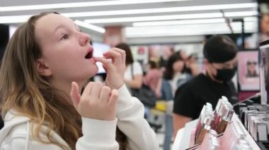 Kozmetik mağazasındaki genç bir kız maskara, ruj, yüz bakımı, kişisel bakım, büyük bir iskele ve arka planda bir sürü insanı seçiyor. Yüksek kalite 4k görüntü