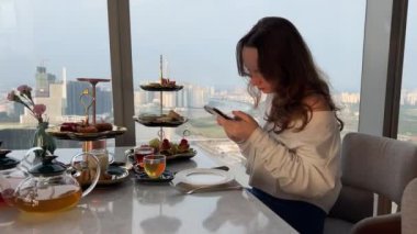 Şık bir restoranda çay partisi, telefonu olan bir kız pastayla çay içiyor ve Ho Chi Minh City Vietnam 'da panoramik bir pencerenin yanında birkaç video çekiyor. Yüksek kalite 4k görüntü