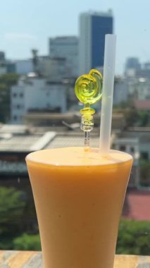Güneşli bir günde, uzun bir bardağa doldurulmuş taze mango suyunun yakın plan görüntüsü. Ho Chi Minh şehrinin arka planında bir restoranda