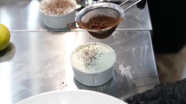 Kadın cam bir kasede tiramisu tatlısı hazırlıyor. Yanında da mutfak kasesi var. Adım adım tarif et. Tiramisu işlem yapıyor. 