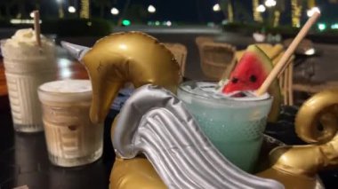 Sahilde limonata bardağında, şişme ördek boynuzlu atıyla sahilde içki içmek. Plaj ışıklarının altında buzlu latte muzlu kremalı karpuz karışımı palmiye ağaçları.