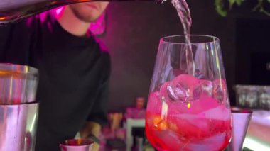 Barda yeşil gömlekli bir barmen şurup, tonik, pembe tatlı bir içecek ve şişeden parlayan büyük bardağa cin döküyor. İçerik sosyal ağları, TV monitörleri. Restoran, otel işi..