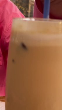 Genç İspanyol kadın güneşli restoran terasında buzlu kahve içiyor. Pembe kazaklı kız sokak kafesinde buzlu latte içiyor.