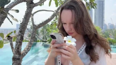 Cep telefonu kullanan bir kadın. Avrupalı iş kadını CEO 'su Fintech uygulamasını kullanarak akıllı telefon tutuyor. Tropik tropikal adaların Plumeria sembolündeki havuzun yanındaki çiçek.