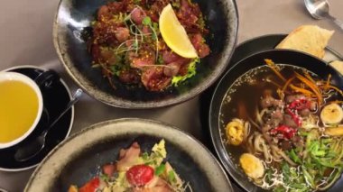 Biftekli salata, barbekü üzümü, Sezar salatası, marul, barbekü hindisi, pastırma ve ton balığı sosu. Erişteli ramen çorbası udon yumurtaları yeşil soğan soya sosu siyah tabakta susam