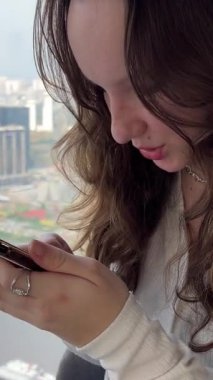 Kadın, akşamları şehir manzarasına bakar. Ho Chi Minh şehrinde gökdelendeki genç kız elinde telefonla oturmuş interneti kullanarak mesaj yazıyor.