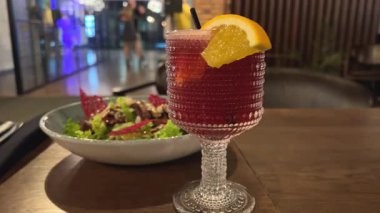 Masada bir kase taze Sezar salatası. Turuncu bardakta kırmızı içki.