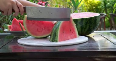 Altın saat boyunca açık havada canlı yeşil kabuklu sulu kırmızı karpuz dilimleyen el yapımı taze, olgun karpuz, yumuşak doğayla çevrili, tabakta parçalara ayrılıyor. Yüksek kalite 4k