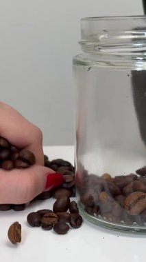 Kırmızı manikürlü bir kız elleriyle bir avuç kahve çekirdeğini alır ve beyaz bir masadaki yeşil bir saksıya, aromatik bir kahve kavanozuna atar. Yüksek kalite 4k görüntü