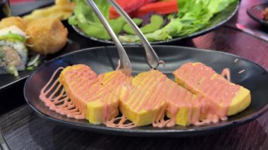 Üzerine sos serpiştirilmiş doğranmış omlet siyah bir tabakta, maşayla, sağlıklı yiyeceklerle, yumurtalarla, salatayla, arka planda suşiyle, Asya 'daki Japon restoranı, açık büfeyle alınır. Yüksek kalite 4k görüntü