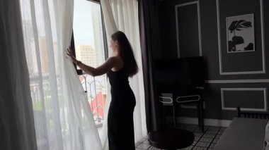 Genç bir kadın perdeleri indiriyor ve geniş pencerenin keyfini çıkarıyor. Yeni güne taze düşüncelerle başlayan bir kız. Yeni başlangıçlar, siyah elbiseli kadın lüks villada perdeleri açıyor.